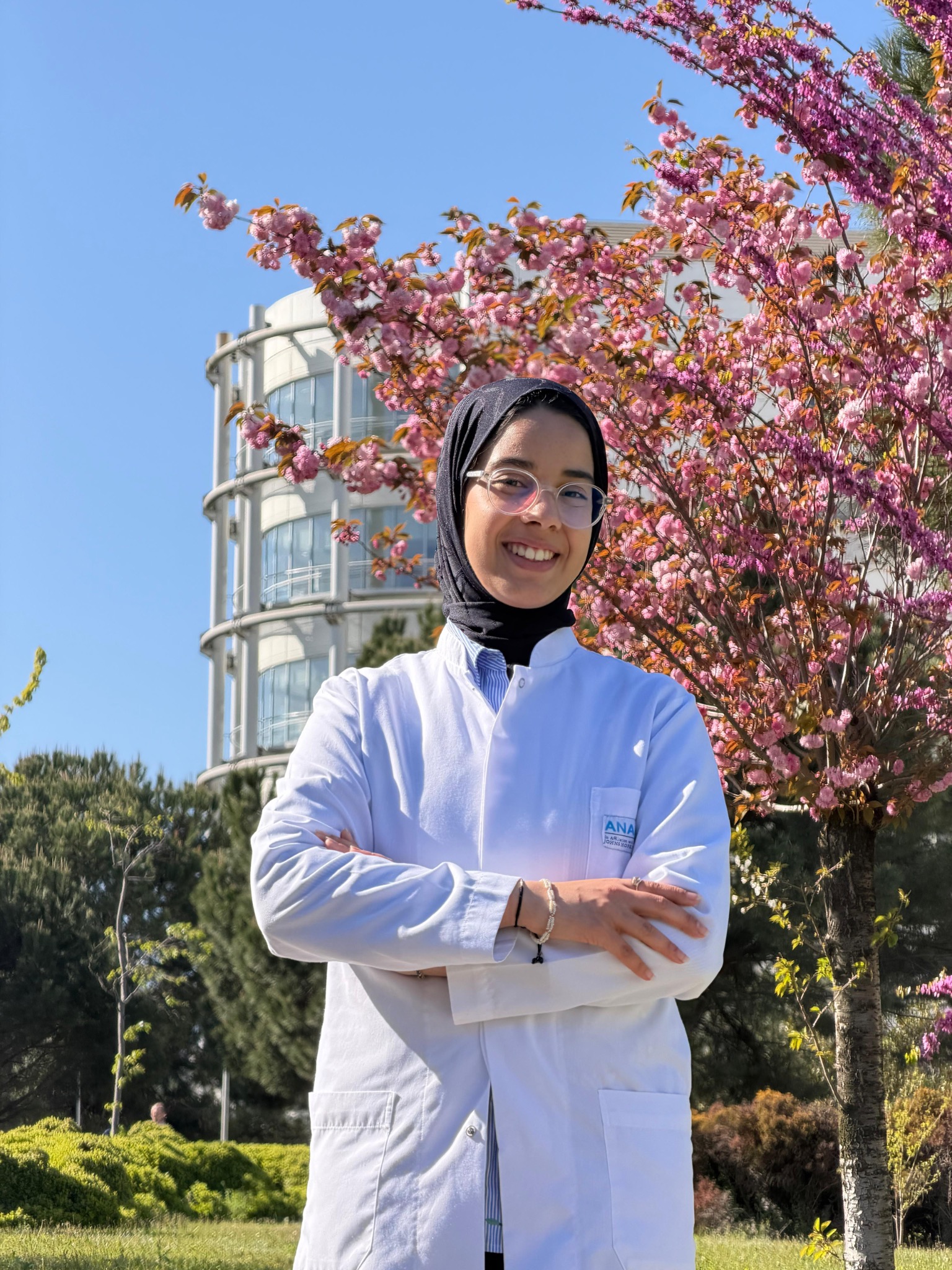 Oumnia smiling in a black hijab and white medical coat beside flowering trees